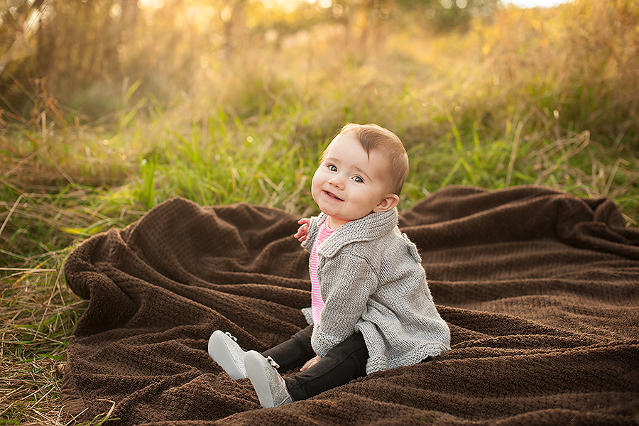 Baby wearing a grey sweater and black pants sitting on a brown blanket