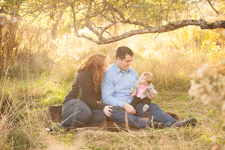 family consisting of a mom and dad, sitting on the ground looking at baby girl in arms