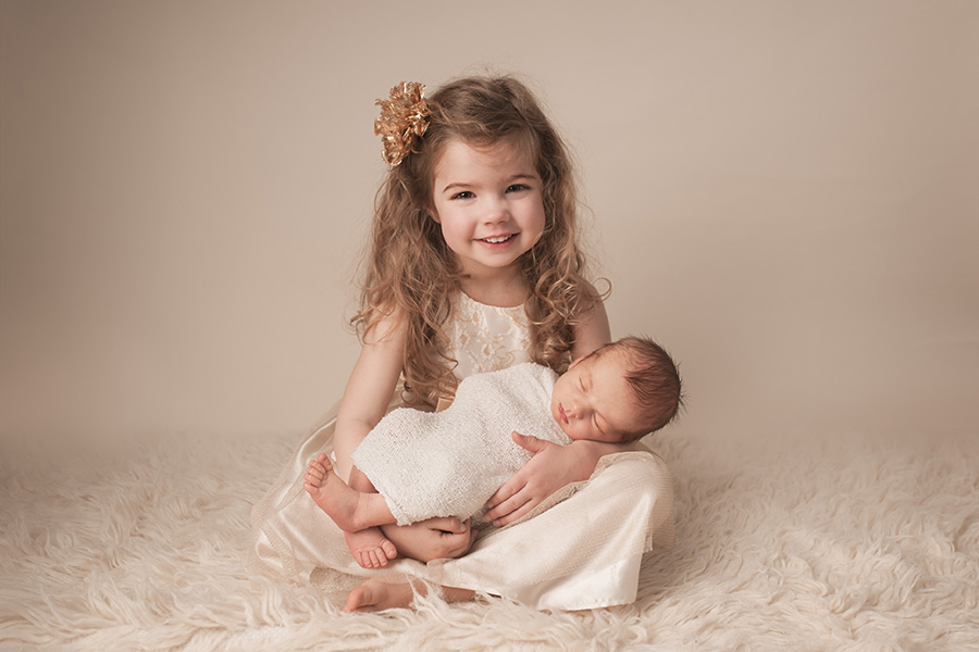 toddler girl in cream dress and curly brown hair smiling while she holds her baby brother 