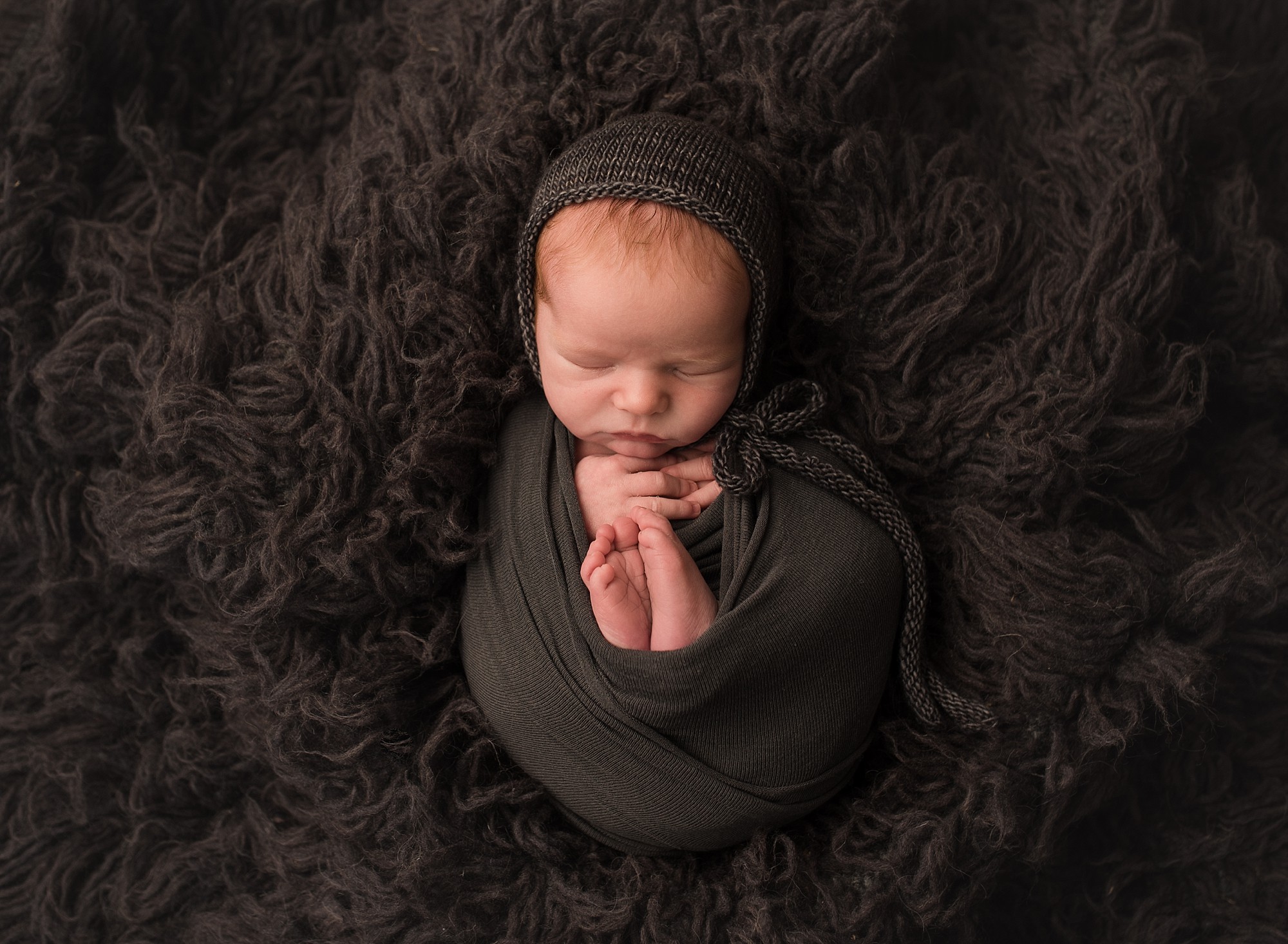 newborn baby wrapped in grey wraps and a grey bonnet and lying on a fire rug