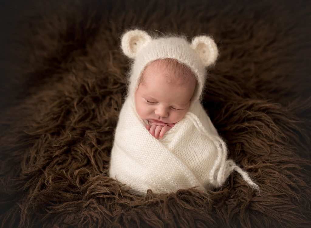 a baby wrapped in a cream wool blanket and wearing a bear hat

