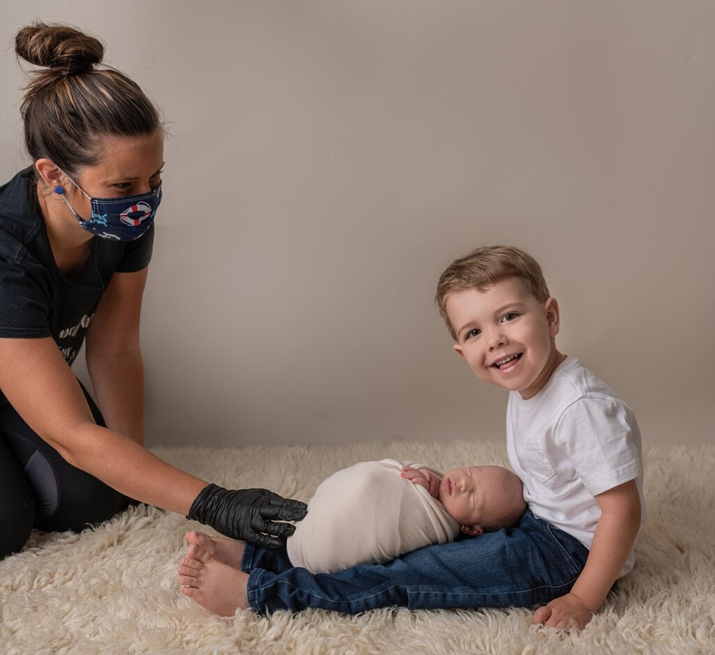 toddler at a photography studio is getting his picture taken and  holding his baby sibling and an adult woman wearing a mask and black plastic gloves is holding the baby and spotting for safety 