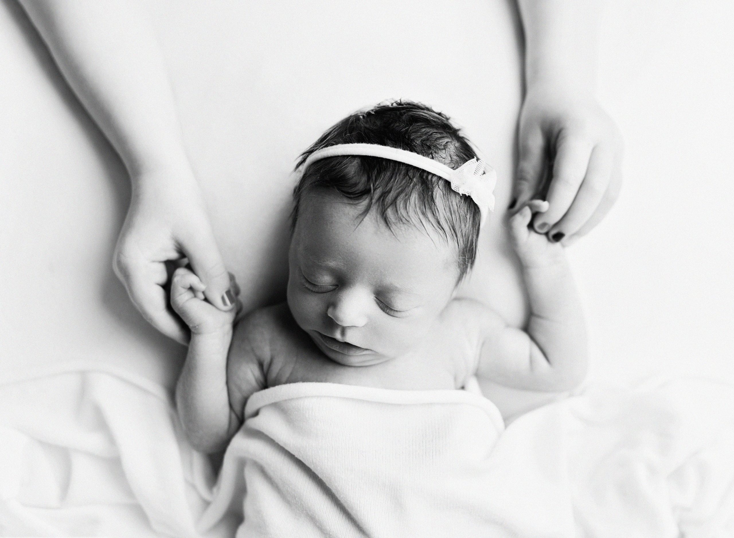 black and white picture of a relaxed posed newborn who is laying on a white blanket and someone is holding their hands