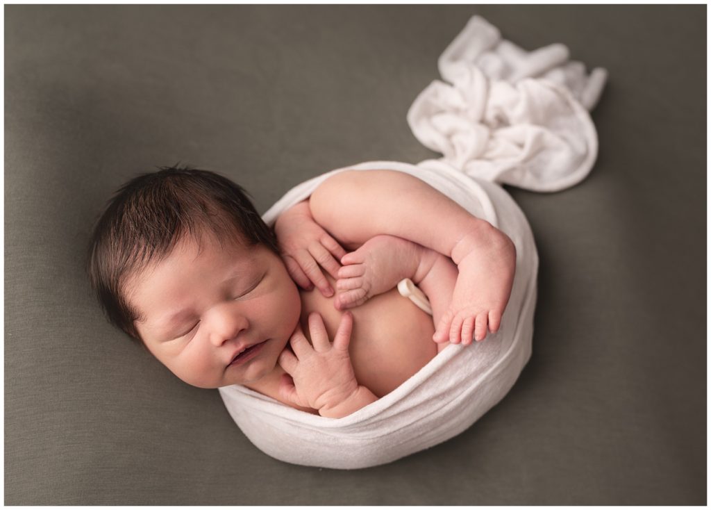 A newborn baby is curled up in a white scarf and sleeping on a smooth blanket at a photo session