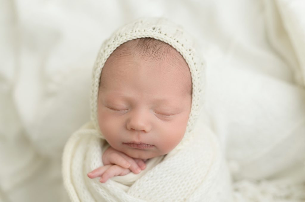 baby sleeping with hands crossed under his chin, wrapped in a knitted blanket and wearing a bonnet.