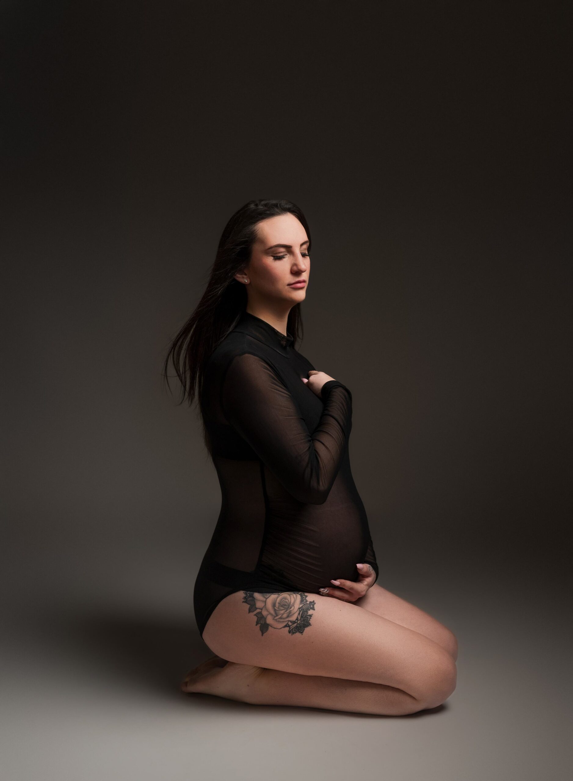 Maternity portrait if a women wearing a black body suit and kneeling on her knees with eyes closed. 