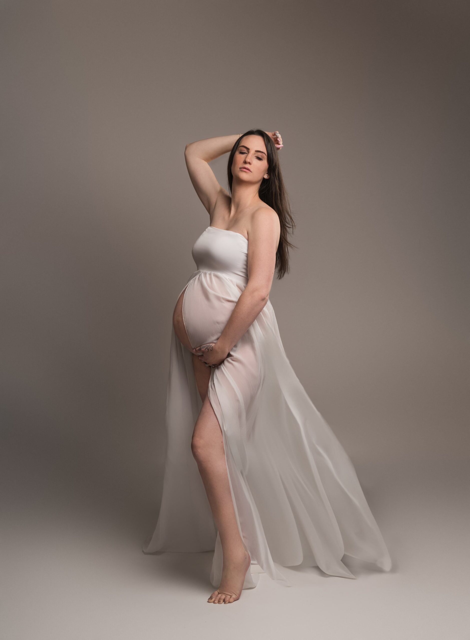 woman posing in a studio for a maternity portraits with her eyes closed wearing a long white gown