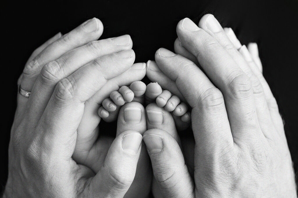 A black and white photo of a larger pair of male hands, cradling smaller female hands which are holding tiny newborn toes.